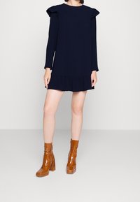 Námornícky modré šaty s dlhým rukávom, riasenými ramenami a vrstveným lemom, skombinované s lesklými hnedými členkovými topánkami s blokovým podpätkom.