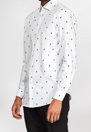 Chemise blanche à manches longues avec boutons, imprimé de sapins verts, portée avec un pantalon noir, visible de la moitié du torse jusqu'au cou et aux mains.