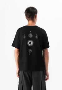 Mann, der ein schwarzes T-Shirt mit weißen Sonnen-, Mond- und Sternsymbolen auf dem Rücken trägt, kombiniert mit dunklen Cargo-Hosen, schaut weg.