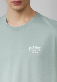 Close-up van een persoon die een lichtgroene t-shirt met ronde hals draagt, met witte tekst die "FNSWHRYR est. Negen Vier GEMAKKELIJKE DAGEN" op de borst zegt.
