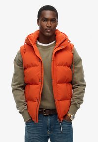 Oranger gefütterter Puffer-Westen mit gepolsterten Bereichen und verstellbarem Kapuzen, getragen über einem grauen Pullover und blauen Jeans. Sichtbare silberne Hardware-Akzente.