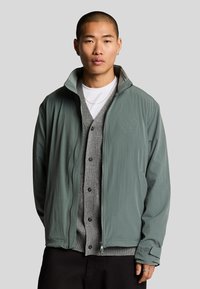 Ljust grön zip-up jacka med ståkrage, framfickor och logotypdetalj. Bärs över en grå kavaj och en vit T-shirt. Slät textur.