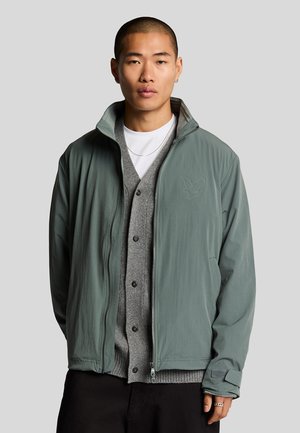 Ljust grön zip-up jacka med ståkrage, framfickor och logotypdetalj. Bärs över en grå kavaj och en vit T-shirt. Slät textur.