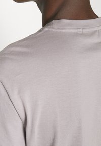 Bagudsigten af en person iført en lys grå T-shirt med rund hals, der viser detaljer omkring nakke, øvre ryg og skuldre.