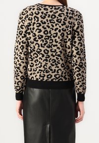 Kobieta w beżowym i czarnym swetrze w doskonały wzór w panterkę, z czarnymi mankietami i dołem, zestawiona z czarną skórzaną spódnicą, pokazana z tyłu.