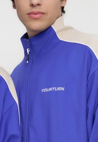 Κοντινό σε άτομο που φοράει μπλε και μπεζ μπουφάν με φερμουάρ και το λέιζερ "YOURTURN" κεντημένο στο στήθος.