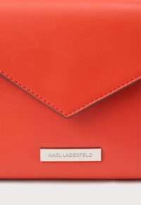 Geantă tip plic din piele roșie, cu o textură netedă, dotată cu o placă de metal cu logo și cusături contrastante pe margini.