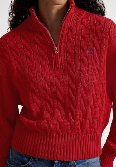 Κόκκινο πουλόβερ με πλέξη καλώδιο, με φερμουάρ μέχρι τη μέση και ριμπ μπάσκα, φορεμένο με μπλε τζιν, εμφάνιση του κορμού και μέρους του λαιμού και των χεριών.