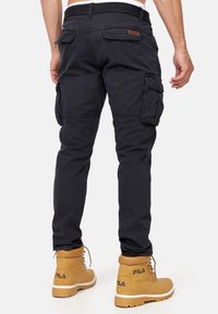Pantalones cargo de algodón en azul marino oscuro con múltiples bolsillos laterales, un acabado texturizado y un diseño ajustado. Llevados con botas color tan.