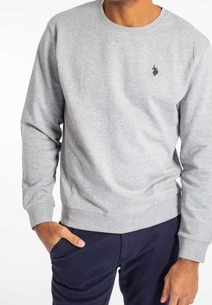 Mann trägt hellgrauen langärmligen Sweatshirt mit kleinem schwarzem Sticklogo auf der Brust und dunkelblauen Hosen, Hand in der Tasche.