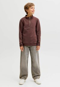 Sweatshirt zippé en polaire marron avec logo en relief, associé à un pantalon en denim gris ample et des baskets blanches. Texture lisse, design décontracté.