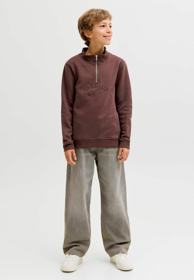 Sweatshirt zippé en polaire marron avec logo en relief, associé à un pantalon en denim gris ample et des baskets blanches. Texture lisse, design décontracté.
