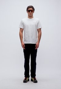 Άνδρας που φοράει λευκό μπλουζάκι Columbia, μαύρο παντελόνι, αθλητικά γυαλιά ηλίου και αθλητικά παπούτσια, στέκεται μπροστά σε απλό λευκό φόντο.