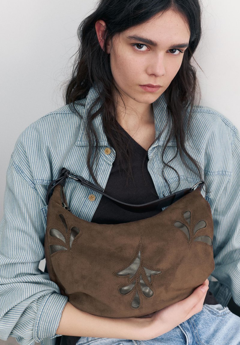 Jeune personne aux longs cheveux foncés portant une veste rayée et une chemise noire, tenant un sac à main en daim marron avec des détails sombres en forme de feuille.