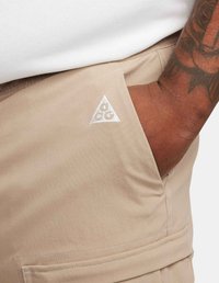 Beiga shorts i lättviktig tyg med sidofickor, med en triangulär vit logotyp och detaljerad sömnad vid fållen.