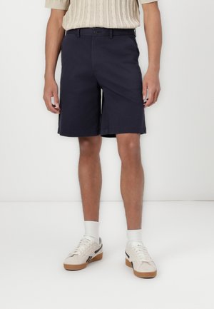 Navy blue shorts made of cotton with a straight fit, featuring a button closure and side pockets. Worn with white sneakers and crew socks.