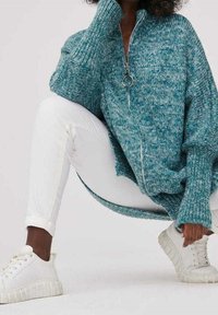 Turkusowy, oversize'owy sweter zapinany na zamek błyskawiczny z teksturą prążkowaną, z metalowym uchwytem, noszony na białych dopasowanych spodniach i białych sneakersach.