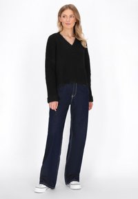 Schwarzer, flauschiger Strickpullover mit V-Ausschnitt, kombiniert mit hoch taillierten, dunkelblauen, weiten Jeans. Weiße Sneaker runden das Outfit ab.