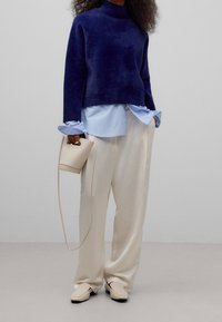 Mørkeblå blød sweater over lyseblå skjorte med knapper, kombineret med cremefarvede bukser med vide ben og beige slingback sko, holder en lille taske.