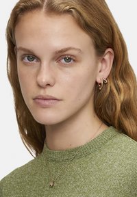 Guldörhängen och ett hängsmycke bärs tillsammans med en grön strukturerad tröja. Håret är vågigt och axellångt, med minimal makeup synlig.