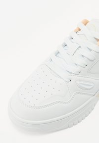 Baskets en cuir blanc avec bout perforé, lacets blancs plats, accents orange clair sur la languette et semelle en caoutchouc texturée.