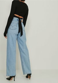 Femme portant un jean large taille haute bleu clair, des bottines noires à talons et un haut court noir à manches longues noué dans le dos, debout sur un sol blanc.