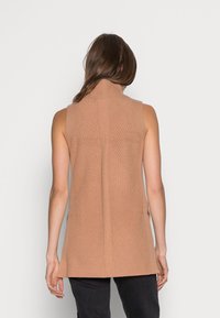 Donna che indossa un gilet in maglia color cammello senza maniche con collo alto, vista da dietro su uno sfondo bianco semplice.