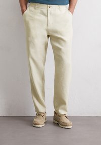 Pantalons en lin beige clair avec une coupe décontractée, dotés de poches latérales et d'une fermeture à bouton, associés à des chaussures en daim marron clair.