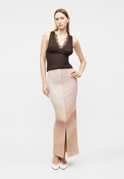 Γυναίκα που φοράει αμάνικο μαύρο τοπ με δαντέλα, μακριά φούστα με κουμπιά σε παστέλ διαβάθμιση και ψηλοτάκουνα λευκά παπούτσια με μυτερή μύτη, στέκεται με το χέρι στη μέση.