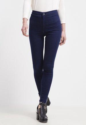 Jeans skinny de tiro alto de mezclilla azul oscuro con una textura suave y sin desgastes visibles; combinados con botines de tacón negro con tiras.