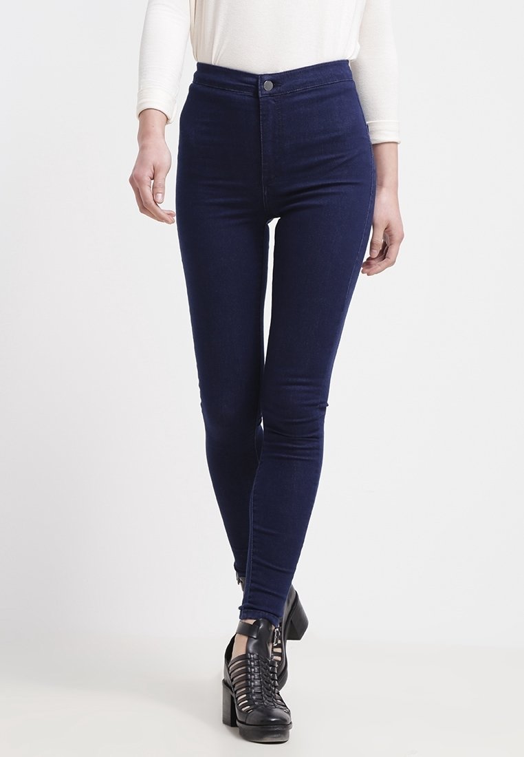 Vysoké kalhoty z tmavě modrého denimu, skinny fit, s hladkou texturou a bez viditelného opotřebení; v kombinaci s černými obuvními botami na podpatku se pásky kolem kotníků.