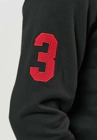 Patch numéro trois rouge cousu sur la partie supérieure de la manche d'un vêtement noir à manches longues.