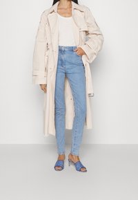 Trench coat color beige chiaro sopra una canotta bianca, abbinato a jeans a vita alta blu chiaro e sandali con tacco blu con un motivo a logo.