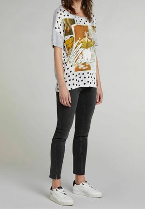 T-shirt blanc à pois noirs avec un col gris, orné d'imprimés graphiques, associé à un jeans slim noir et des baskets blanches.