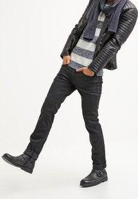 Giubbotto di pelle nera con accenti a costine, maglione grigio a righe, jeans neri e stivaletti con dettaglio a cinturino. La modella assume una posa casual.