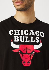 Melns kokvilnas T-krekls ar lielu sarkanu Čikāgas Bulls logo, kurā attēlots vērša galva, un taupīgu baltu tekstu "CHICAGO BULLS".