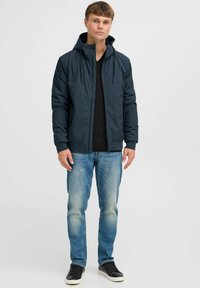 Chaqueta navy con cremallera y capucha, puños elásticos, combinada con jeans azul claro y zapatillas negras. Diseño simple y funcional.