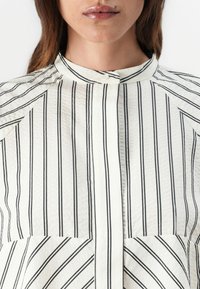 Vit skjorta med tunna svarta vertikala ränder, hög krage, knappstängning, textil med struktur och två framfickor.