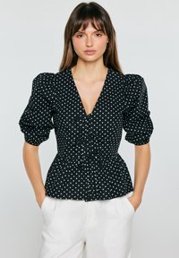 Mujer con blusa negra de mangas abullonadas con lunares blancos y detalle de lazo, combinada con pantalones blancos de tiro alto, manos en los bolsillos.