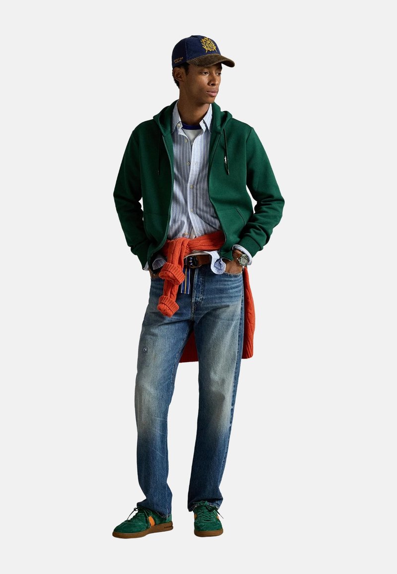 Modelo vistiendo un sudadera con capucha verde sobre una camiseta a rayas azul, con un suéter naranja atado a la cintura, jeans azul claro y zapatillas verdes.