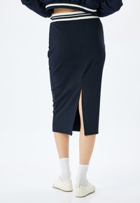 Jupe crayon bleu marine avec une fente haute, fabriquée en tissu doux, dotée d'une taille rayée blanche contrastante. Portée avec des chaussures et des chaussettes blanches.