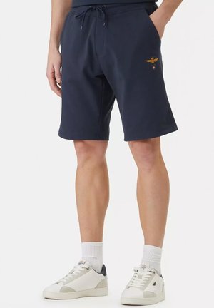 Uomo che indossa pantaloncini blu navy con coulisse e emblema dorato ricamato sulla coscia, calzini bianchi e sneakers basse bianche.