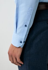 Camicia azzurro chiaro con un motivo testurizzato, con polsini con bottoni e bottoni blu scuro. Indossata con pantaloni scuri, mostra un design aderente.