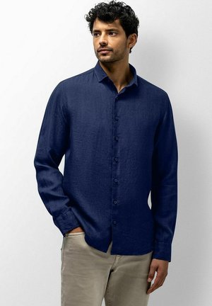 Man met donker krullend haar, die een donkerblauw overhemd met lange mouwen en beige broek draagt, staand tegen een lichte achtergrond.