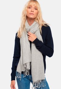 Femme blonde portant un pull marine et un jean bleu, tenant une écharpe frangée gris clair enroulée autour de son cou.
