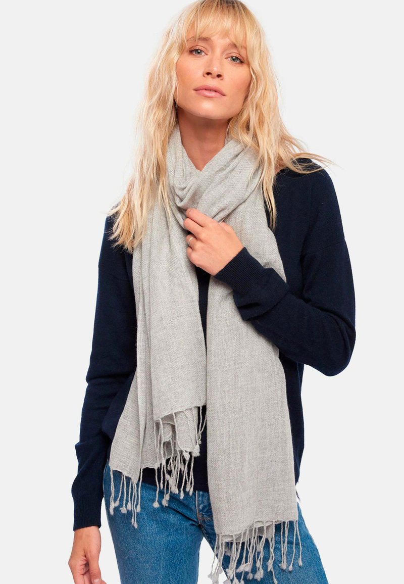 Femme blonde portant un pull marine et un jean bleu, tenant une écharpe frangée gris clair enroulée autour de son cou.