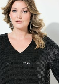Top negro con lentejuelas y escote en V, que presenta un diseño texturizado y mangas drapeadas. La modelo lleva pendientes cuadrados adornados con rhinestones.