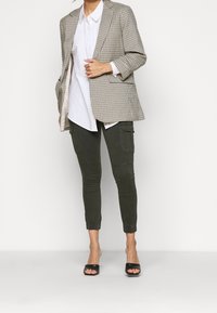 Pruhovaný blazer v béžové a hnědé barvě, pod ním bílá košile, olivově zelené cargo kalhoty a černé klouzavé sandály. Slim-fit s ohrnutými kotníky.