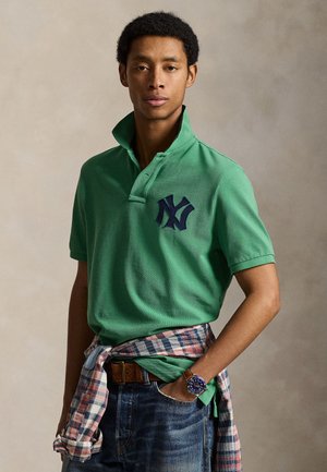 Mladý muž v zelenej polo košeli s tmavým logom, kockovanej košeli previazanou okolo pása, modrých džínsoch a s náramkovými hodinkami, stojaci s rukou v vrecku.