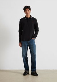 Schwarzes Langarm-Polohemd mit einer Dreiknopf-Leiste, versehen mit einem kleinen Logo, kombiniert mit blauen geraden Jeans und schwarzen Schuhen.